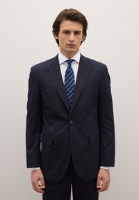 Navy blue, two-button suit with a notch lapel and side pockets, worn over a white dress shirt, paired with a blue striped tie.