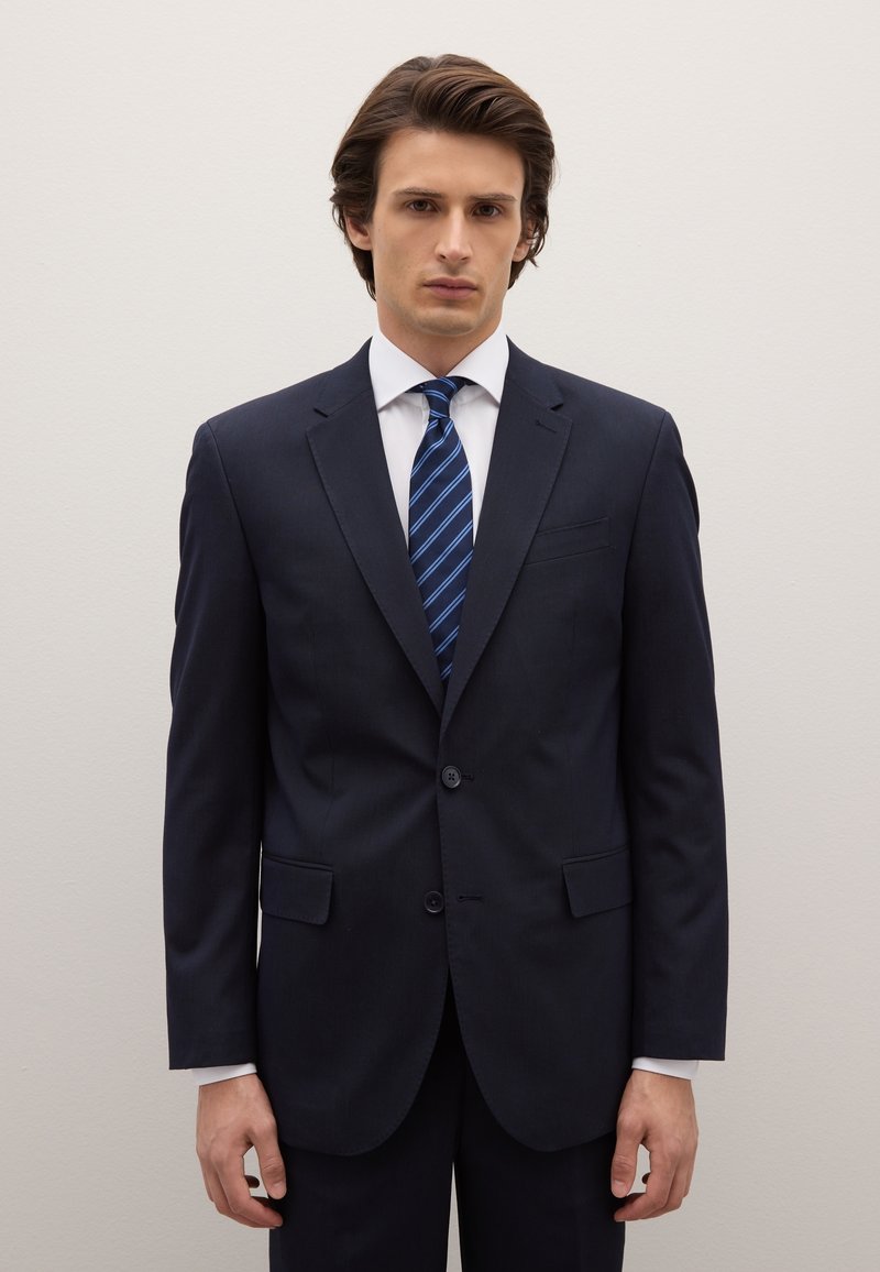 Navy blue, two-button suit with a notch lapel and side pockets, worn over a white dress shirt, paired with a blue striped tie.