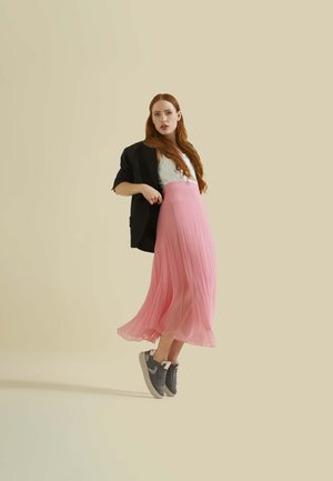 Jupe plissée rose clair, blazer noir et baskets grises, sur un fond neutre. Le tissu est fluide avec une texture douce.