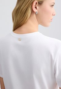 Hvid bomulds t-shirt med korte ærmer, der har en lille cirkulær metallisk detalje på ryggen. Blød tekstur og afslappet pasform.