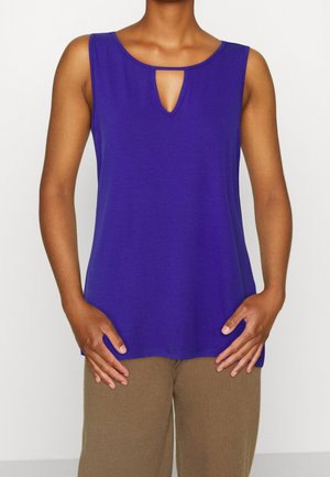 Fioletowy top bez rękawów z zaokrąglonym dekoltem i małym wycięciem w kształcie łezki z przodu. Gładka faktura, luźny krój, w połączeniu z beżowymi spodniami.