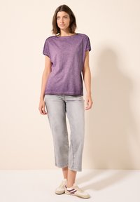 Haut violet à manches courtes avec une coupe décontractée, doté de détails en boutons sur les épaules, associé à un jean cropped gris clair et des baskets beiges.