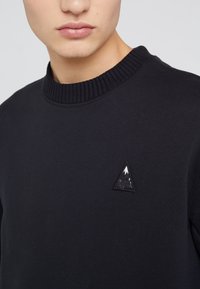 Sudadera negra con cuello de canalé, que presenta un pequeño logotipo de triángulo bordado en blanco y negro en la parte delantera. Textura suave y lisa.