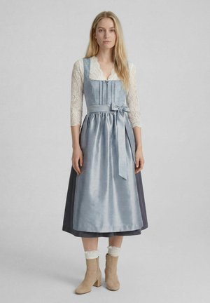 Junge Frau mit langen blonden Haaren trägt ein blaues traditionelles Kleid mit Spitzenärmeln, eine an der Taille gebundene Satin-Schürze, weiße Knöchelsocken und beige Stiefel.