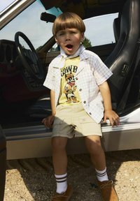 Bambino che indossa una maglietta grafica gialla, una camicia a maniche corte bianca a quadretti, pantaloni beige, scarpe marroni e calzini bianchi con righe marroni, seduto in un'auto.