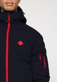 Marinblå isolerad jacka med röd dragkedja och detaljer, med hög krage och logopatch på bröstet samt fackdetalj på ärmen.