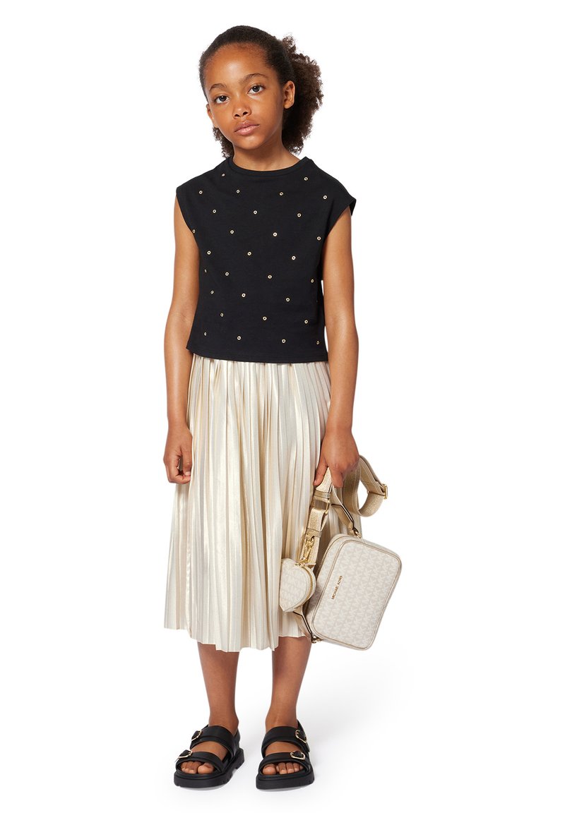 Haut noir à manches courtes avec des clous dorés, jupe midi plissée métallique, et sandales noires. L'enfant porte un sac à bandoulière beige à motifs.
