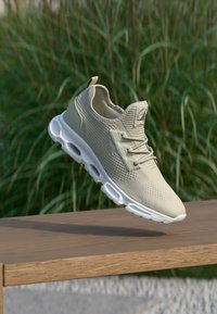 Hellgrauer Sneaker mit strukturiertem Textilobermaterial, auffälligem Schnürsystem, weißer, gepolsterter Sohle und abgerundeter Form, der auf einer Holzoberfläche ruht.