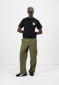 Black t-shirt with a circular green logo, olive cargo pants with side pockets, and a gray/green patterned cap. Black sneakers complete the outfit.