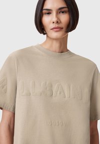 T-shirt in cotone beige con scollo rotondo, caratterizzata da un logo "ALLSAINTS" in rilievo e testo "LONDON" in una finitura liscia. Maniche corte, vestibilità rilassata.