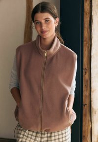 Hellrosa Fleece-Weste mit hohem Kragen, Reißverschluss auf der Vorderseite und seitlichen Taschen, getragen über einem grauen Langarmoberteil und gemusterten Karohosen.