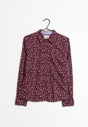 Camisa burdeos de manga larga con botones y pequeño estampado blanco similar a plumas, colgada en una percha negra sobre fondo blanco.