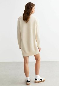 Crème-kleurige oversized sweaterjurk met geribbelde zoom en manchetten, gemaakt van zacht materiaal, gedragen met stijlvolle sneakers en witte sokken.