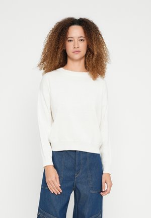 Άσπρο πλεκτό πουλόβερ με στρογγυλή λαιμόκοψη και μακριά μανίκια, με άνετη εφαρμογή. Φορεμένο με σκούρο τζιν παντελόνι, που παρουσιάζει ένα απλό σχέδιο.