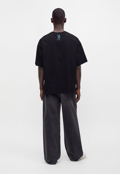 Man wearing black oversized t-shirt with small blue astronaut graphic on upper back, loose dark gray pants, and white shoes, standing facing away.