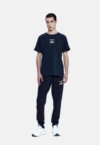 T-shirt in cotone blu navy e joggers abbinati, con branding bianco sul petto e sulla coscia, abbinati a scarpe sportive bianche.