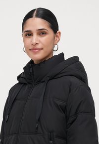 Mujer con cabello oscuro en una parte central, usando aretes de aro plateados y una chaqueta de plumas negra con capucha y cordones.