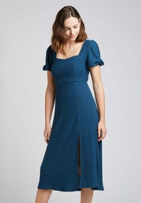 Robe bleue texturée avec des manches bouffantes, décolleté carré et fente sur le côté, présentant un corsage ajusté et une jupe fluide, en tissu extensible.