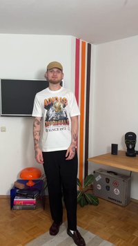 Balti Adidas marškinėliai su grafiniu dizainu ir tekstu, juodos kelnės ir rudi batai. Kambarys su oranžine lempa, augalais ir spalvingomis sienų juostelėmis.