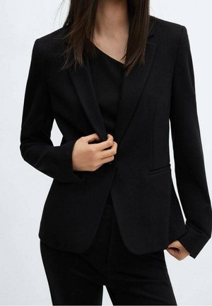 Kvinde iført en tætsiddende sort blazer og bukser, justerer blazeren med den ene hånd mod en ensartet lys baggrund.