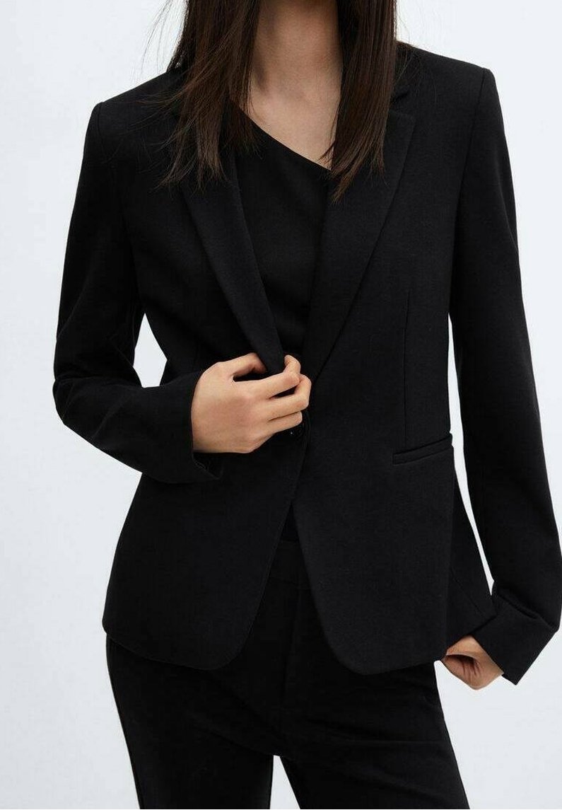 Femme portant un blazer noir ajusté et un pantalon, ajustant le blazer d'une main, sur un fond clair uni.