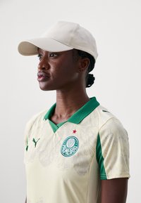 Bāla dzeltena polo krekls ar zaļo apkakli, Puma logs un Palmeiras emblēma. Komplektā ar gaiši bēšu beisbola cepuri.