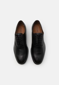 Chaussures habillées noires en cuir avec une finition lisse, bout rond et lacets fins. L'intérieur présente une semelle intérieure marron clair avec un logo.