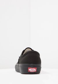 Zapatilla de lona negra con un diseño de perfil bajo, suela de goma y una etiqueta roja distintiva "Vans Off The Wall" en el talón.