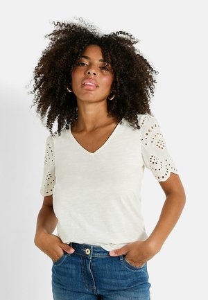 Camiseta blanca de cuello en V con mangas cortas de encaje, combinada con jeans azules. La tela tiene una textura sutil y un corte relajado.