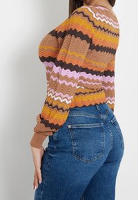 Sweter dzianinowy w zygzakowy wzór w odcieniach brązu, pomarańczu, różu i żółci, w połączeniu z niebieskimi dżinsami o wysokim stanie. Długie rękawy z ściągaczami.