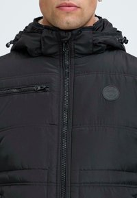 Black padded jacket with a zip closure, high collar, and adjustable hood; features a chest pocket and a round logo patch on the front.