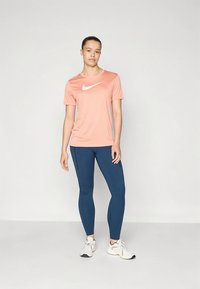 Világos rózsaszín Nike póló fehér logóval, sötétkék leggingsszel és fehér sportcipővel párosítva. A ruházat kényelmes, hétköznapi kialakítású.