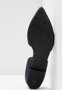 Chaussure en tissu bleu foncé avec un bout pointu et une semelle en caoutchouc. La semelle présente des pièces de monnaie en relief et du texte avec un logo, ainsi qu'une surface texturée.