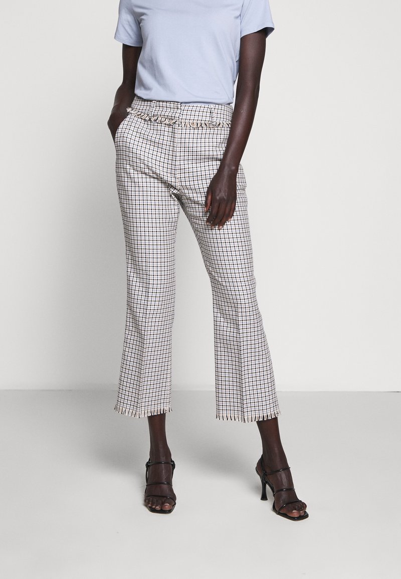 Femme portant un pantalon court à carreaux avec des ourlets à franges, un haut bleu clair à manches courtes et des sandales noires à talons, debout devant un fond uni.