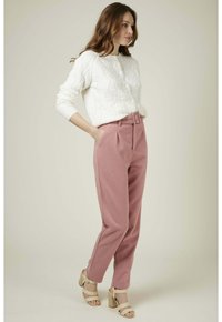 Pull en maille blanche avec des boutons ronds, associé à un pantalon rose taille haute avec une ceinture. Le mannequin porte des talons épais de couleur claire.