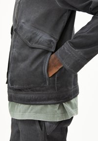 Schwarze Baumwolljacke mit Fronttaschen, strukturierte Gestaltung und glatte Textur, über eines hellgrünen Hemd getragen.