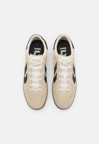 Beżowe zamszowe sneakersy z czarnymi akcentami w zygzak, białymi sznurówkami i czarną gumową podeszwą. Logo widoczne na języku i pięcie.