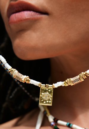 Collier doré avec des perles blanches, des détails métalliques raffinés et un pendentif rectangulaire avec un design en forme d'œil ressemblant à un diamant.