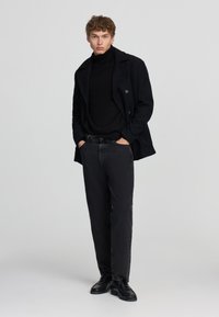 Giovane uomo in piedi con le mani nelle tasche, vestito con un cappotto nero, un dolcevita nero, jeans scuri e scarpe nere, su uno sfondo chiaro e uniforme.