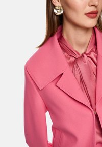 Rosa taillierter Blazer mit breitem Kragen, strukturierten Schultern und glatter Textur; geschichtet mit einem satinrosa Schal. Goldene runde Ohrringe.