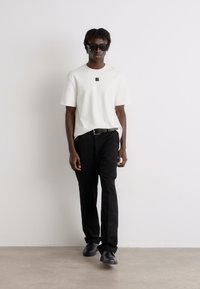 Weißes T-Shirt mit kurzen Ärmeln und kleinem Logo, kombiniert mit schwarzen Hosen und schwarzen Schuhen. Das Model trägt dunkle Sonnenbrillen.