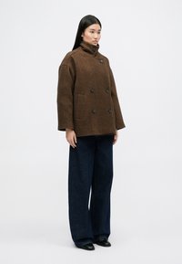 Καφέ παλτό με σχέδιο ρομβοειδούς, υψηλό κολάρο, διπλό κούμπωμα και δύο μπροστινές τσέπες, συνδυασμένο με φαρδιά σκούρα τζιν και μαύρα παπούτσια.