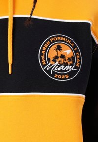 Gaubtas megztinis su oranžiniais, juodais ir baltais skydeliais. Savo centre turi siuvinėtą McLaren logotipą su žodžiais „Miami“ ir „2025“. 