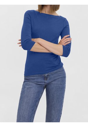 Kobieta ubrana w niebieską bluzkę z rękawami do łokcia i niebieskie dżinsy, stojąca z założonymi ramionami na tle gładkiej białej ściany.