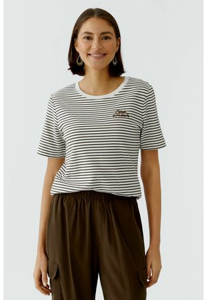 Frau lächelnd, trägt ein schwarz-weiß gestreiftes Kurzarmshirt mit Text und dunkelbraune Hose mit Taschen.