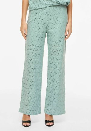 Femme portant un pantalon taille haute à jambes larges de couleur vert menthe avec un motif en tissu ajouré et des sandales à talons ouvertes noires.