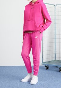 Pink Nike hoodie and matching sweatpants, made of soft fabric, featuring a front pocket, drawstrings, and logo accents. Worn with white sneakers.