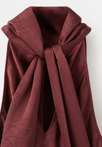 Bordeauxroter Stoff, locker drapiert und zu einem Knoten gebunden, mit subtiler Textur und Falten, vor einem neutralen Hintergrund.