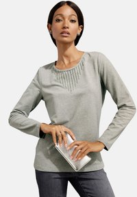 Helles graues Langarmshirt mit einem mit Perlen verzierten Ausschnitt, kombiniert mit einer silbernen Clutch. Das Model steht mit einer Hand auf der Clutch und präsentiert das Outfit.