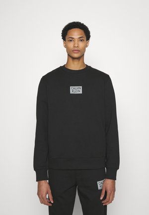 Felpa nera con collo rotondo e etichetta grigia rettangolare che presenta "CALVIN KLEIN" in bianco, tessuto testurizzato, e polsini ed orlo elastico.
